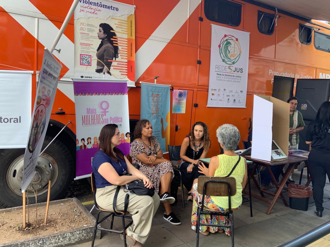 Três mulheres sentadas conversam com uma idosa em frente a um ônibus laranja com cartazes sobre enfrentamento à violência.