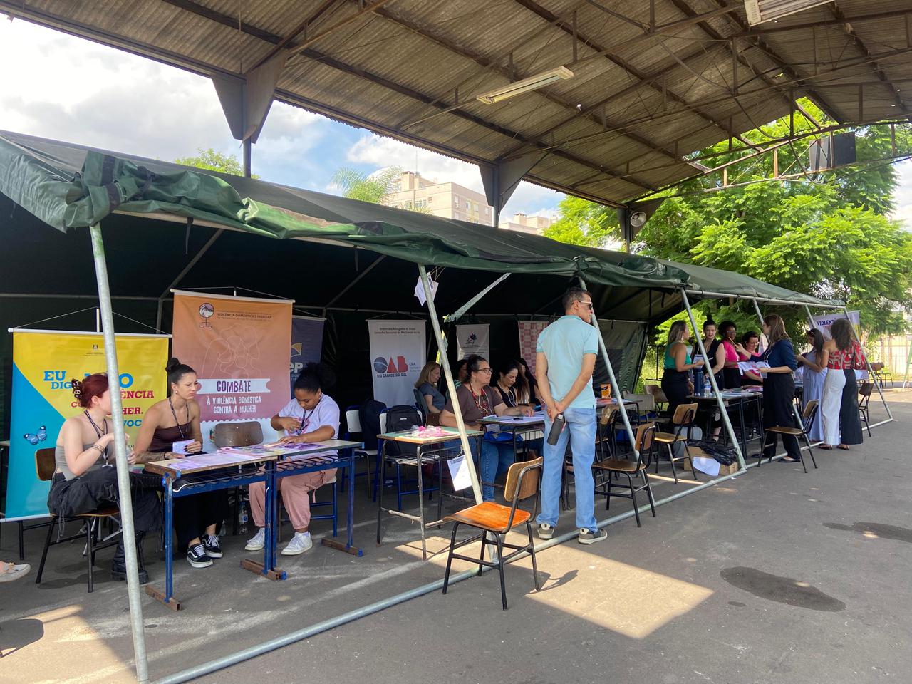 Tenda com mesas de atendimento onde mulheres prestam orientações ao público em ação sobre direitos e combate à violência.