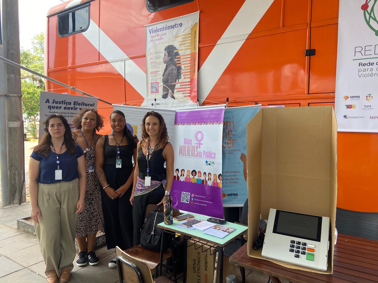 Quatro mulheres posam em frente a um ônibus laranja, com banners sobre direitos das mulheres e participação política.