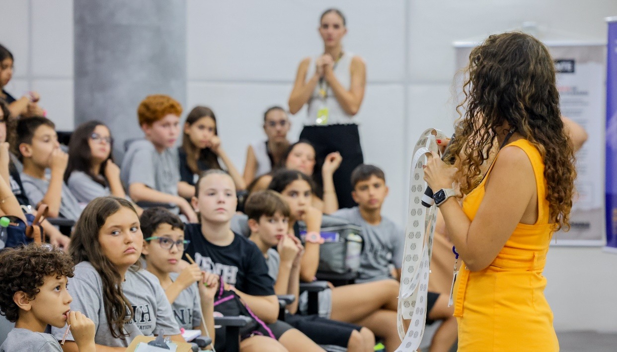 uma mulher com vestido amarelo, ela está com os votos da urna nas mãos, enquanto fala o resultado no microfone.
