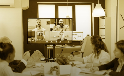 A imagem ilustra uma mesa de reuniões com pessoas sentadas em uma videoconferência com colegas de trabalho.